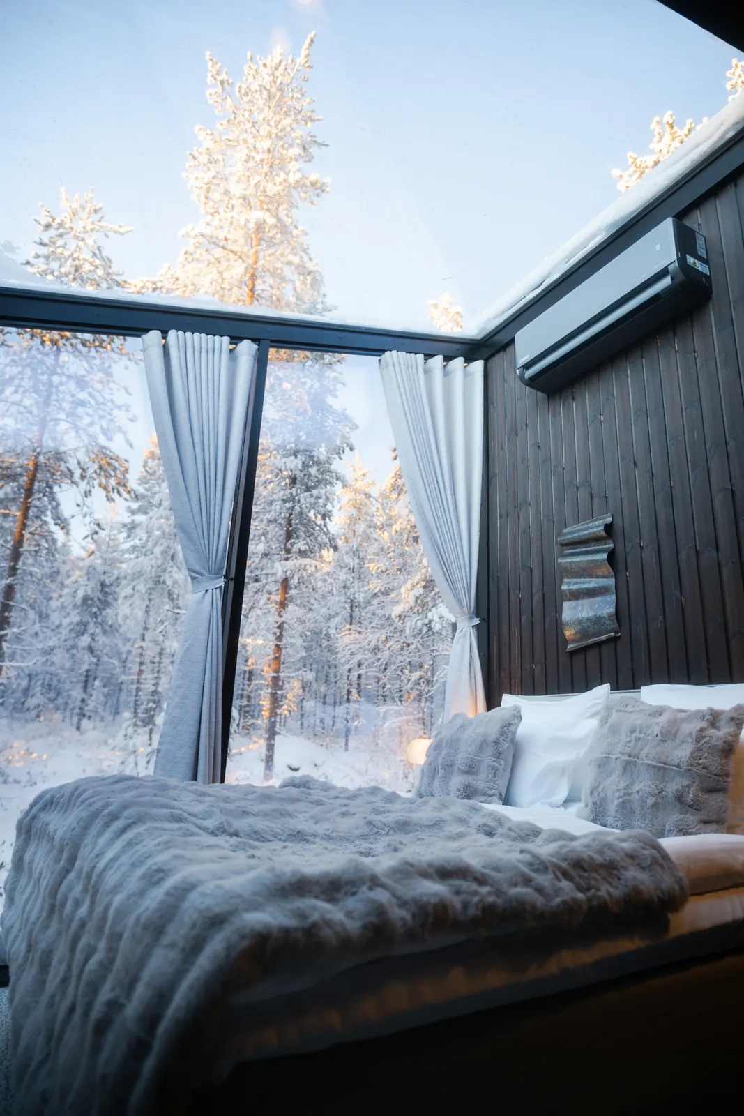 Inside a Hillside igloo: a double bed with a fur throw beside a glass wall looking onto snowy forest