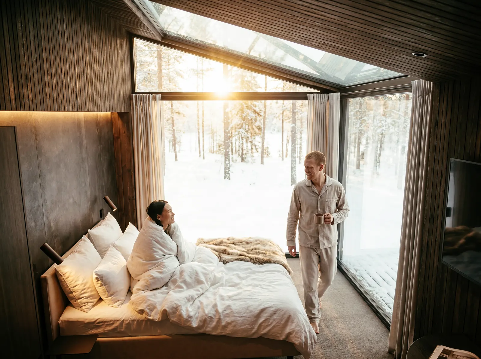 A couple in a Riverside igloo with a glass roof, the bed facing tall windows onto snowy pines